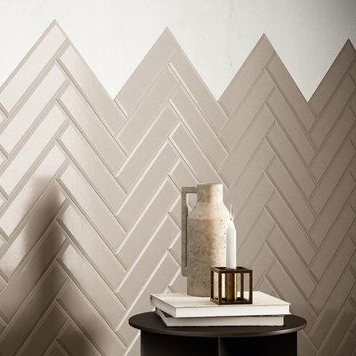 Mur intérieur décoratif en céramique beige posée en chevrons avec finition mate, mettant en valeur un style contemporain minimaliste, accentué par une table d’appoint noire sur laquelle reposent un vase en béton, des livres et une bougie décorative moderne par Déco Surfaces.