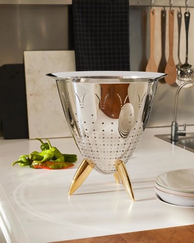 Passoire design en acier inoxydable avec motifs décoratifs et pieds dorés signée Max le Chinois par Philippe Starck pour Alessi, posée sur un comptoir de cuisine moderne avec légumes frais et accessoires en arrière-plan offert chez Boutique Crème