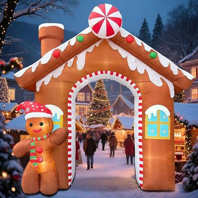 Arche gonflable de Noël en forme de maison pain d'épices avec bonhomme en pain d'épices souriant, installée dans un village de Noël illuminé avec un grand sapin décoré à l'arrière-plan par AMAZON