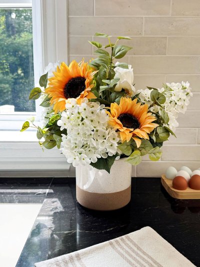 Arrangement floral avec tournesols, fleurs blanches et feuillage vert dans un vase bicolore, posé sur un comptoir de cuisine près d'une fenêtre ensoleillée. par Décors Véronneau