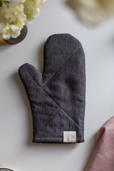Mitaine de four en lin anthracite fait main par Confetti Mill, posée sur une surface blanche avec un coin de nappe rose et un bouquet de fleurs en arrière-plan – produit localement à Montréal offert par Boutique Crème