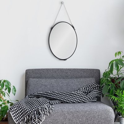 Miroir rond avec cadre noir de chez Lovaco suspendu au-dessus d’un canapé gris décoré d’un jeté à pois noirs et blancs, entouré de plantes vertes