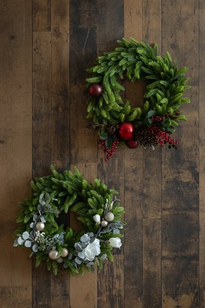 Deux couronnes de Noël décoratives sur un plancher de bois, l'une ornée de boules rouges, de pommes de pin et de baies, l'autre avec des fleurs blanches, feuillage givré et boules dorées - Décors Véronneau