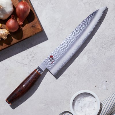 Couteau de chef Gyutoh au fini martelé avec manche en bois foncé de Eugène Allard, posé sur un plan de travail près d’ingrédients frais