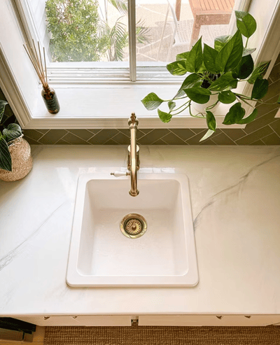 Évier de cuisine blanc encastré dans un comptoir en quartz à effet marbré, muni d’un robinet doré au style vintage, placé devant une fenêtre laissant entrer une lumière naturelle abondante, dosseret en tuiles vert olive en chevron et plantes décoratives apportant une touche de fraîcheur organique de chez Muraluxe
