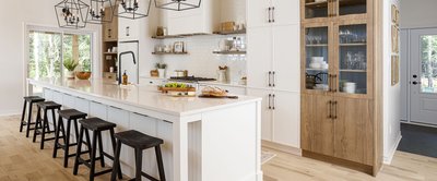 Cuisine de style farmhouse avec un long îlot blanc et tabourets noirs, armoires en bois naturel et blanc, dosseret en céramique blanche, étagères ouvertes et luminaires suspendus en métal noir au-dessus de l’îlot. Design signé Armoires Cuisines Action