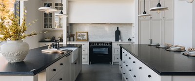 Cuisine élégante au style champêtre moderne avec armoires crème, deux îlots avec comptoirs noirs contrastants, évier de ferme, robinetterie noire, luminaires suspendus au design industriel et cuisinière noire de style classique. Design signé Armoires Cuisines Action
