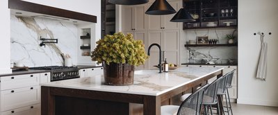 Cuisine haut de gamme avec comptoir et dosseret en marbre veinuré, armoires crème et noires, îlot central en bois foncé avec évier moderne noir mat, et luminaires coniques suspendus, par Armoires Cuisines Action