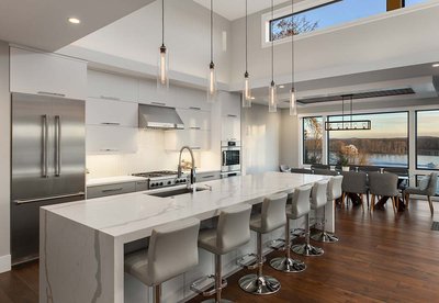 Cuisine moderne par Rénovilla avec armoires blanches sans poignées, grand îlot en quartz veiné, chaises hautes grises, électroménagers en acier inoxydable et vue panoramique sur un lac grâce à de grandes fenêtres pleine hauteur