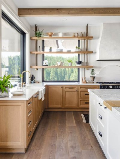 Cuisine lumineuse de style champêtre moderne signée Onivo, avec armoires en bois beige, comptoirs en quartz blanc, évier sous plan, étagères ouvertes devant une grande fenêtre donnant sur la forêt