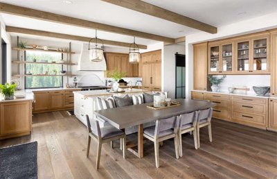 Cuisine champêtre moderne signée Onivo, avec armoires en bois beige clair, poutres apparentes, grand îlot central blanc avec tabourets, table à manger intégrée pour huit personnes et vue panoramique sur la forêt