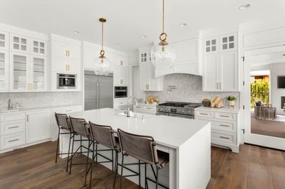 Cuisine blanche de style contemporain signée Onivo, avec armoires shaker, comptoir en quartz blanc, électroménagers encastrés en inox, dosseret en mosaïque de marbre, luminaires suspendus en verre et plancher en bois foncé