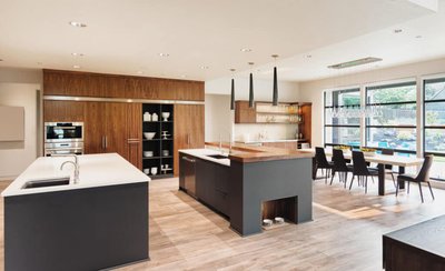 Cuisine moderne signée Onivo avec armoires en noyer, deux îlots aux comptoirs contrastants en quartz blanc et bois, électroménagers encastrés en inox, coin repas avec grandes fenêtres pleine hauteur et chaises noires élégantes