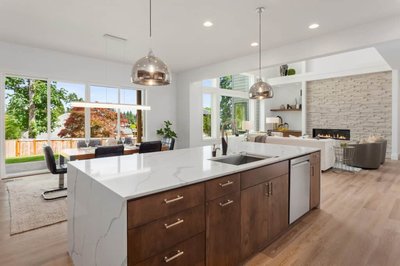 Cuisine ouverte de style contemporain signée Onivo avec îlot central en bois noyer et comptoir en quartz blanc à veinures grises, luminaires suspendus chromés, vue sur salle à manger lumineuse et salon avec foyer mural en pierres claires