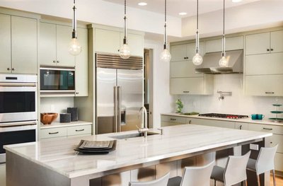 Cuisine moderne par Onivo avec armoires vert pâle, électroménagers en acier inoxydable intégrés, grand îlot central en quartz veiné, luminaires suspendus à ampoules visibles et chaises de comptoir contemporaines