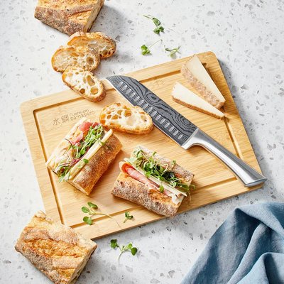 Couteau Santoku Damashiro au design damassé et manche métallique, présenté sur une planche en bois de Eugène Allard avec sandwichs, fromage et tranches de pain