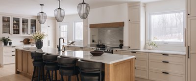 Cuisine lumineuse au style classique et contemporain avec armoires crème, comptoirs en quartz pâle, îlot central en bois avec tabourets noirs, luminaires suspendus en forme de cage et grande fenêtre laissant entrer la lumière naturelle. Design signé Armoires Cuisines Action