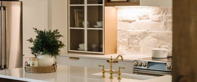 Cuisine chaleureuse et contemporaine avec armoires blanches, comptoir en quartz lustré, robinetterie dorée de style vintage, dosseret en pierre naturelle texturée et décor de Noël minimaliste. Design par Armoires Cuisines Action