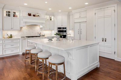 Cuisine classique blanche signée Rénovilla avec armoires moulurées, grand îlot central avec comptoir en quartz, tabourets en bois rembourrés, électroménagers intégrés et plancher de bois franc chaleureux