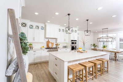Cuisine contemporaine signée Rénovilla avec armoires blanches vitrées, grand îlot central en quartz, tabourets en bois, luminaires suspendus noirs et planchers en bois clair – ambiance lumineuse et conviviale