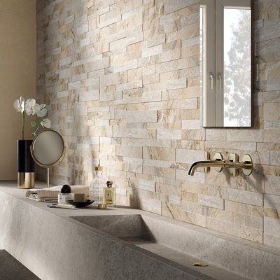 Salle de bain élégante avec revêtement mural en pierre naturelle aux tons beiges, robinetterie murale dorée, lavabo en béton moulé et décor minimaliste incluant un miroir circulaire et des accessoires de luxe par Déco Surfaces.