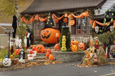 Maison décorée pour l'Halloween avec de nombreuses citrouilles, un grand Jack-o'-lantern avec chapeau de sorcière, des fantômes, des épouvantails et des guirlandes orange.