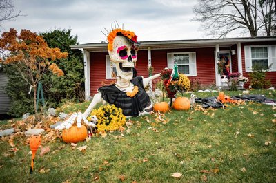 Cour avant décorée avec un grand squelette habillé de noir et orné de fleurs colorées, entouré de citrouilles, de chrysanthèmes jaunes et d'éléments festifs inspirés du Dia de los Muertos.