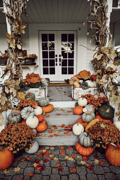 Balcon décoré pour l'Halloween avec des citrouilles orange, blanches et vertes, des pots de chrysanthèmes, des feuilles colorées et des tiges de maïs séchées devant une porte blanche.