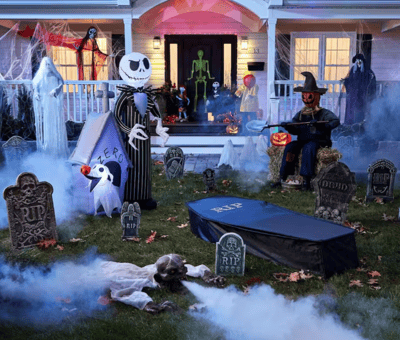 Cour avant décorée pour l'Halloween avec pierres tombales, personnages effrayants, squelette, citrouilles, effets de brouillard et lumières colorées devant une maison illuminée.