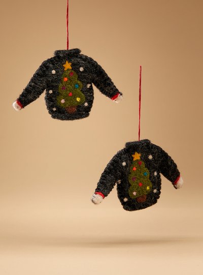 Deux petites décorations en forme de pulls en laine suspendus par des fils rouges. Les pulls sont noirs avec un motif de sapin de Noël décoré de boules colorées et surmonté d'une étoile jaune. Des petits points blancs imitent des flocons de neige sur le fond beige.