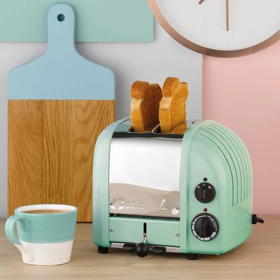 Grille-pain 2 tranches Dualit de couleur vert menthe, au design rétro, avec deux rôties dorées, placé sur un comptoir blanc accompagné d'une tasse de café et de planches à découper pastel, dans une cuisine au style moderne et coloré offert chez Boutique Crème
