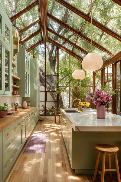 Cuisine lumineuse avec armoires vert sauge, comptoirs en bois et marbre, îlot central, grandes fenêtres et plafond en verre donnant sur un jardin arboré Source - Elevé Living Pinterest