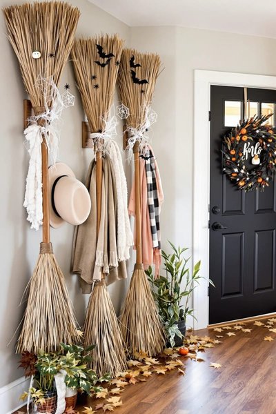 Entrée de maison décorée pour l'Halloween avec des balais de sorcière accrochés au mur, des écharpes, une couronne automnale sur une porte noire et des feuilles d'automne au sol.