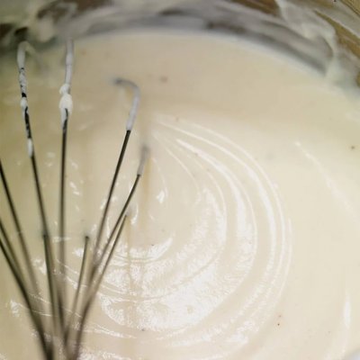 Fouet en silicone de marque Ricardo en train de mélanger une pâte crémeuse de couleur beige dans un bol – ustensile de cuisine polyvalent pour sauces, crèmes et préparations pâtissières, disponible chez La Guilde Culinaire