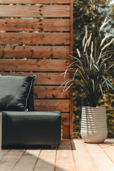 Coin terrasse avec fauteuil noir à coussin contrastant, panneau de bois vertical et grande plante ornementale dans un pot texturé blanc, sur un plancher en bois clair par Décors Véronneau