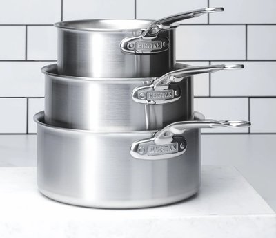 Ensemble de trois casseroles en acier inoxydable Hestan de la collection Thomas Keller Insignia, empilées sur un comptoir en marbre devant un mur à carreaux blancs Offert chez Boutique Crème