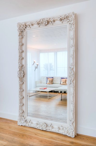 Grand miroir blanc orné de motifs sculptés reflétant un salon moderne lumineux avec canapé blanc et plancher en bois