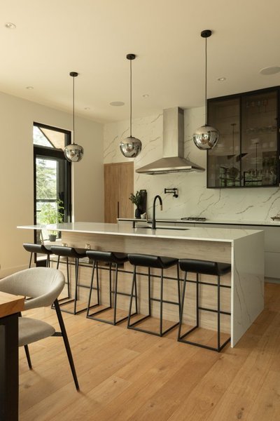 Cuisine contemporaine conçue par Ateliers Jacob avec un îlot en quartz blanc effet marbre, cinq tabourets noirs au design épuré, armoires en bois clair, hotte en inox et luminaires sphériques suspendus, le tout dans un espace lumineux avec plancher de bois franc
