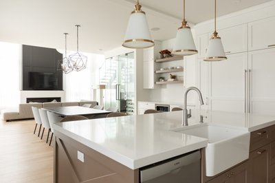 Cuisine moderne et lumineuse signée Ateliers Jacob, avec îlot central brun taupe à évier de ferme blanc, comptoirs en quartz blanc, armoires murales blanches avec poignées dorées, et vue ouverte sur la salle à manger et salon contemporain avec murs vitrés et mobilier minimaliste