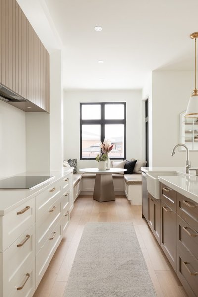 Cuisine lumineuse signée Ateliers Jacob avec armoires blanc cassé et poignées dorées, îlot central avec évier de ferme, coin repas intégré sous une grande fenêtre noire et banquettes rembourrées, sol en bois pâle et ambiance contemporaine chaleureuse