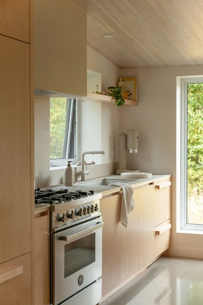 Cuisine compacte et lumineuse de style scandinave conçue par Ateliers Jacob, avec armoires en bois blond, comptoir pâle, cuisinière au fini inox, robinetterie blanche, grande fenêtre laissant entrer la lumière naturelle, et étagère flottante décorée de plantes