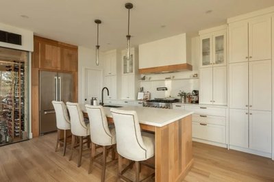Cuisine lumineuse signée Ateliers Jacob avec armoires blanches de style shaker, grand îlot central en bois de chêne blanc avec comptoir pâle, chaises rembourrées élégantes, hotte décorative en bois, électroménagers encastrés en inox et cellier vitré intégré