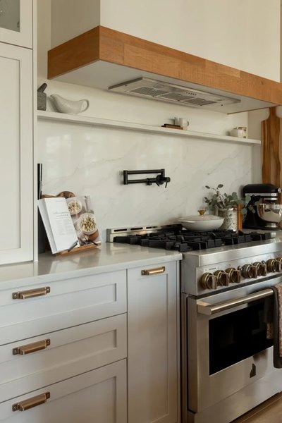 Gros plan sur une cuisinière au gaz en acier inoxydable dans une cuisine signée Ateliers Jacob, avec hotte en bois de chêne blanc, dosseret en céramique effet marbre, robinet mural noir mat, comptoir en pierre claire et armoires shaker blanches avec poignées dorées