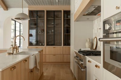 Cuisine moderne haut de gamme signée Ateliers Jacob avec armoires en bois clair, comptoirs en quartz blanc, robinetterie dorée, grand garde-manger vitré noir, cuisinière encastrée, et design aux lignes épurées inspiré du style scandinave