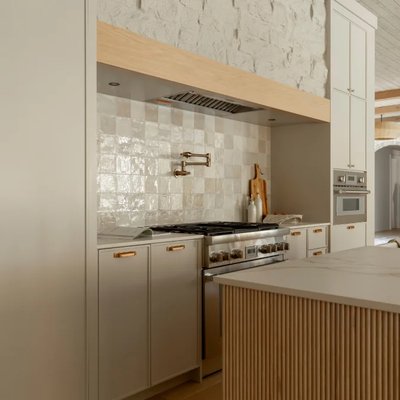Vue rapprochée d'une cuisine signée Ateliers Jacob, avec cuisinière professionnelle en acier inoxydable, armoires crème à poignées dorées, dosseret en céramique artisanale blanche à relief, hotte intégrée sous boiserie claire et électroménagers encastrés