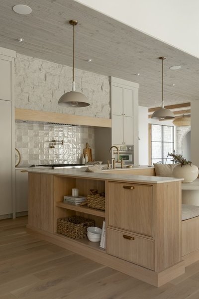 Cuisine haut de gamme signée Ateliers Jacob, avec îlot central en bois clair comprenant des étagères ouvertes et des paniers tressés, armoires murales crème, dosseret en carreaux de céramique texturés blancs, robinets dorés et suspension au style contemporain