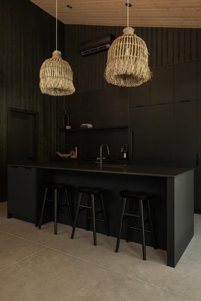 Cuisine contemporaine Ébène conçue par Ateliers Jacob, avec armoires noires pleine hauteur, îlot central épuré et quatre tabourets noirs, mise en valeur par deux luminaires suspendus en fibres naturelles et un plafond en bois clair