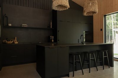 Cuisine moderne Ébène signée Ateliers Jacob avec armoires noires mates, îlot central spacieux et tabourets noirs, éclairée par des suspensions en rotin et une grande fenêtre donnant sur la forêt