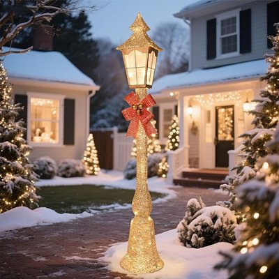 Lampadaire de Noël lumineux décoré d'un nœud rouge pailleté au centre d'une cour enneigée, entouré d'arbustes et de sapins illuminés devant une maison décorée pour les fêtes - Amazon
