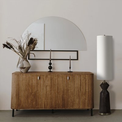 Lampe sur pied design avec base en bois sombre sculptée et abat-jour cylindrique blanc, placée à côté d’un buffet en bois nervuré surmonté d’un vase décoratif et de chandeliers noirs, devant un mur beige avec miroir demi-lune, image provenant de Linen Chest